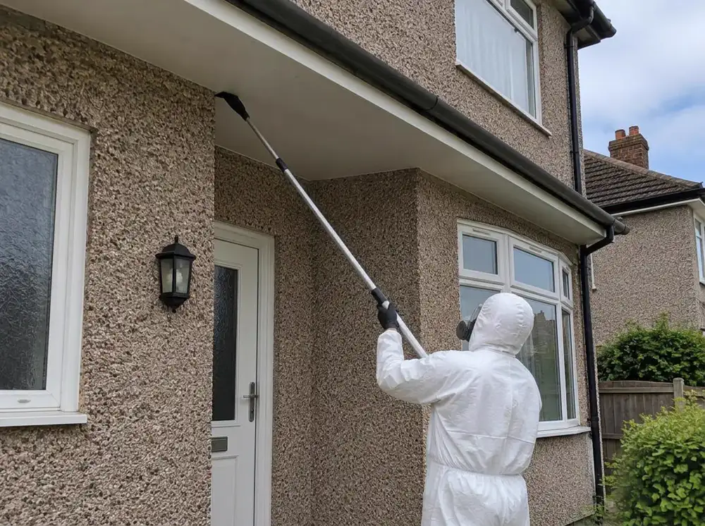 Technician in a white protective suit treating a wasp nest in the corner of a soffit from the ground with a long telescopic lance, at a pebbledash 1930s semi